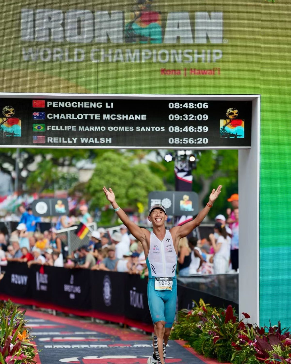 巴斯（李鵬程）在 2026 Kona IRONMAN 世界錦標賽終點拱門下張開雙手衝線，上方計分板顯示 PENGCHENG LI 08:48:06