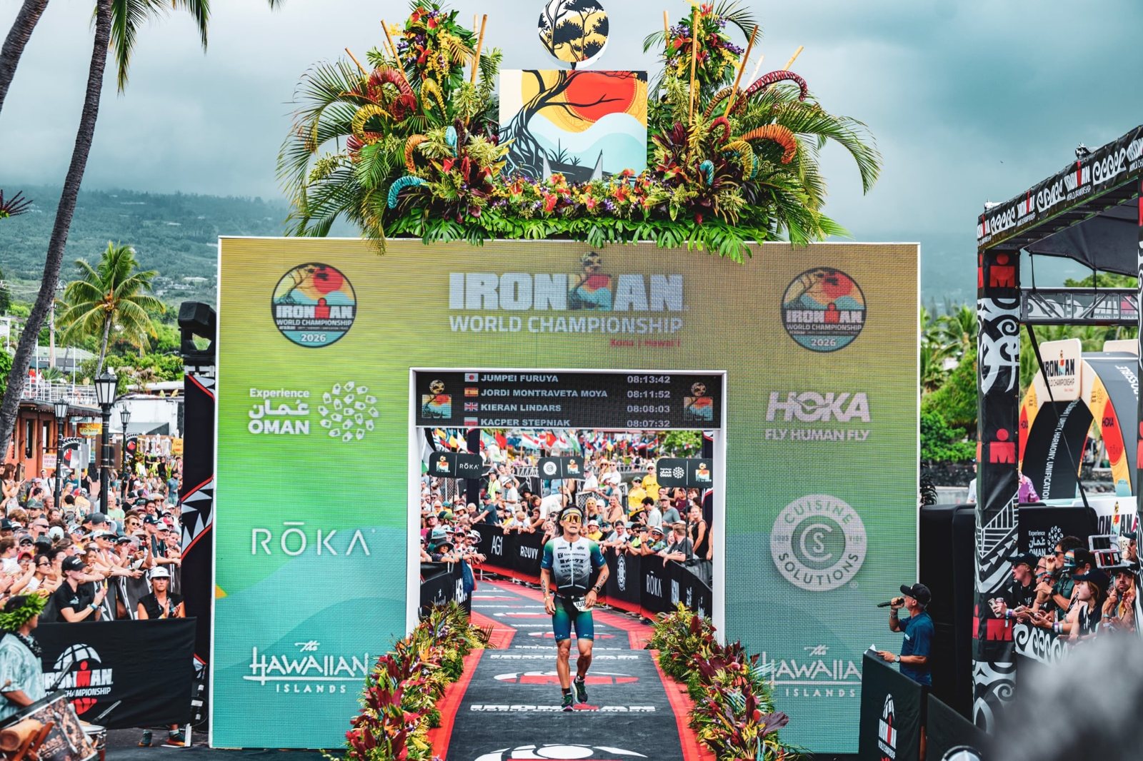 古谷純平在 2026 Kona 終點拱門下，計分板最上面一行顯示 JUMPEI FURUYA 08:13:42