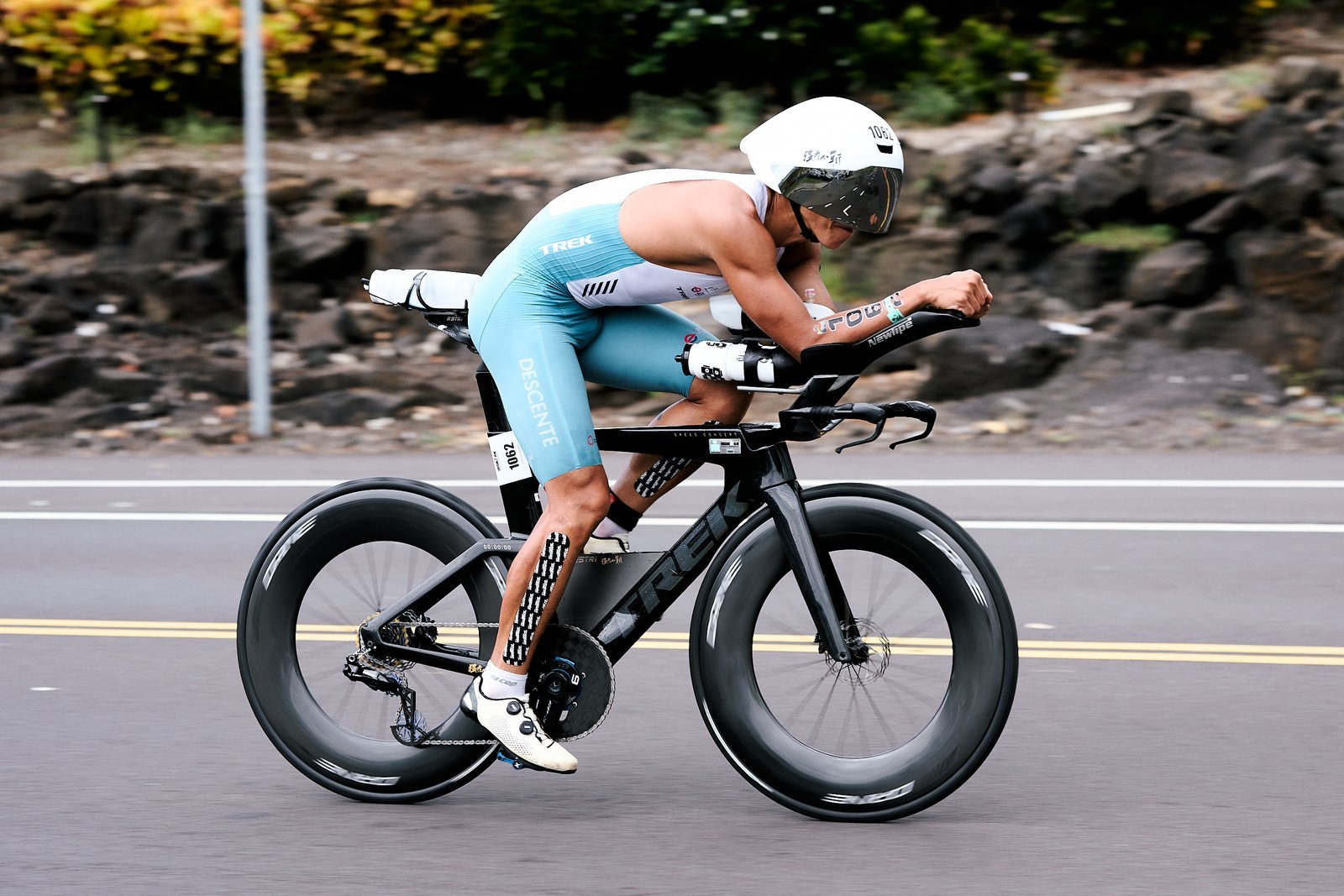 巴斯（號碼 1062）在 2026 Kona IRONMAN 世界錦標賽騎車段趴在計時把手上，背景是熔岩石牆